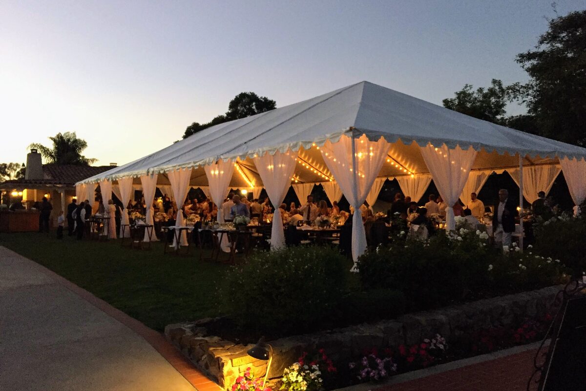 An evening reception under a white marquee strung with lights, guests gathered at tables on a lawn