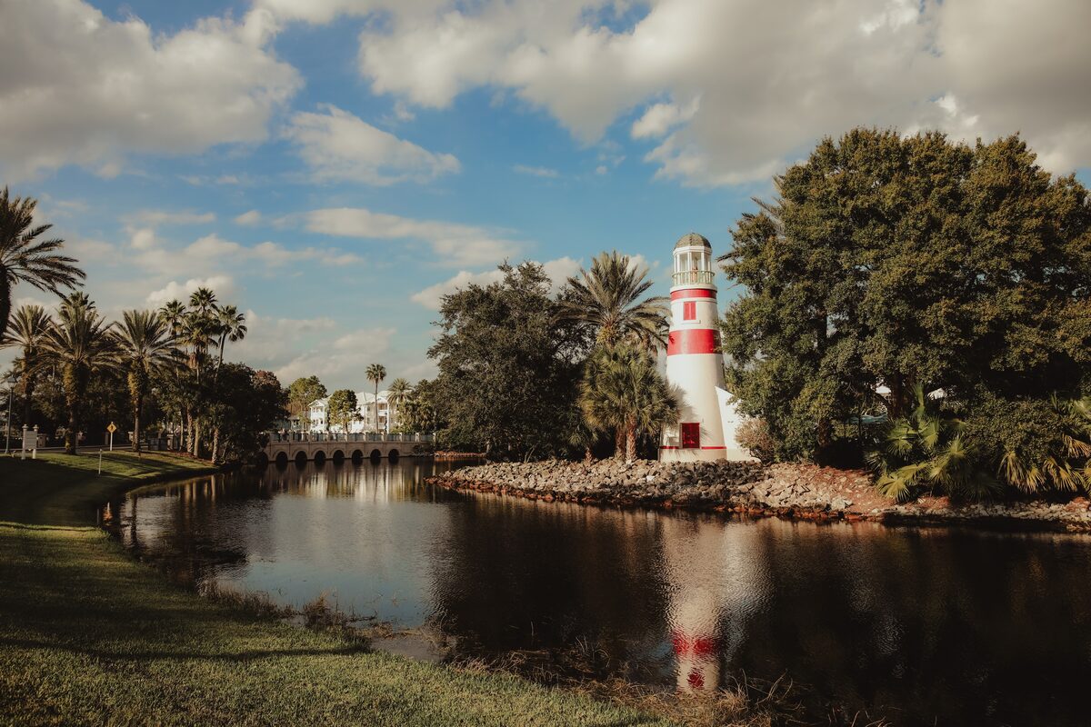 A small red-and-white lighthouse beside a palm-lined lake at a resort, with a stone bridge in the distance